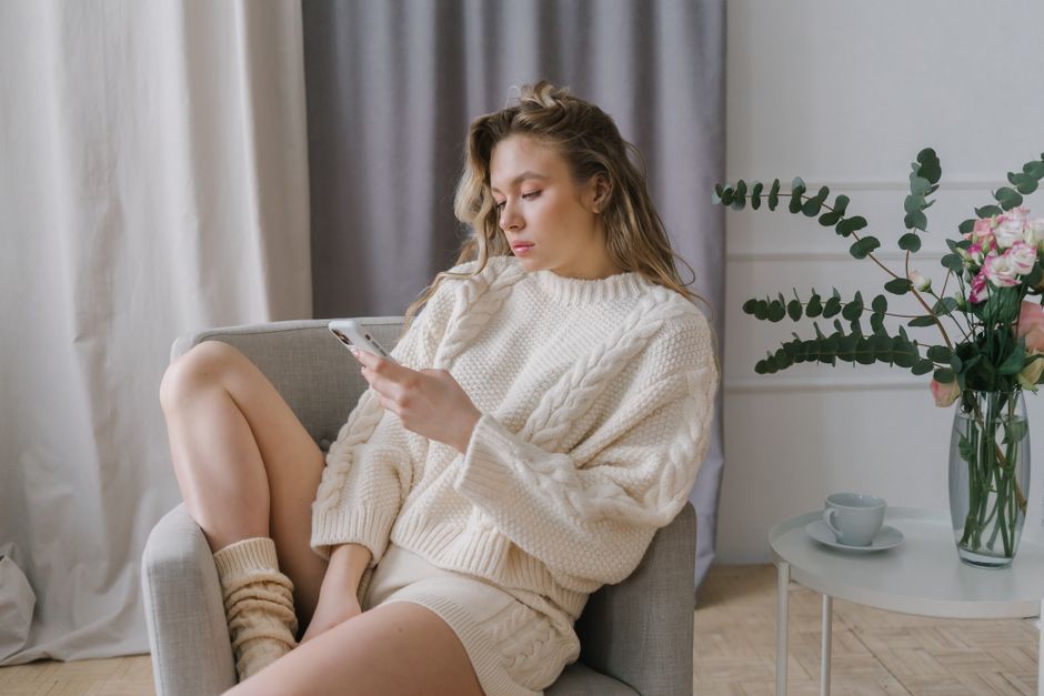Woman in cozy sweater using smartphone in a modern indoor setting, relaxing on an armchair